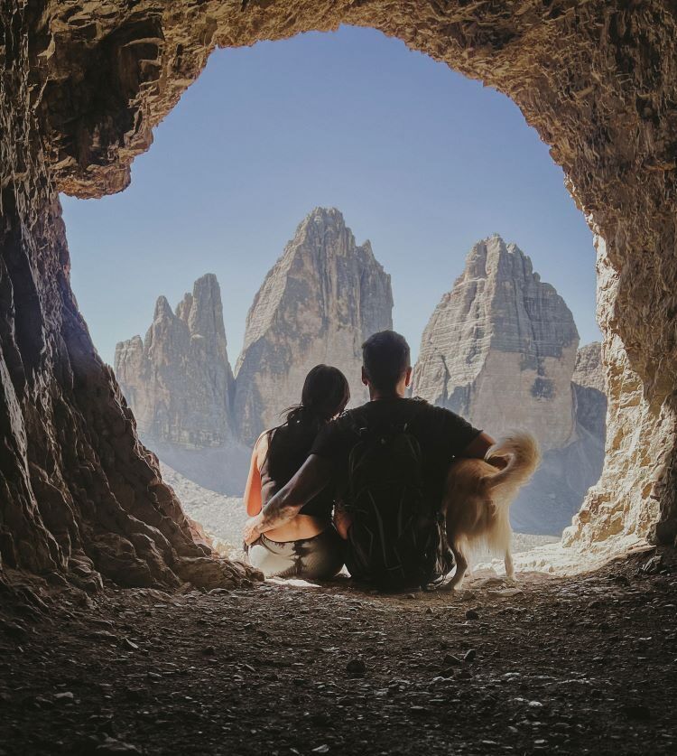 cueva y pareja sentada mirando a las montañas abrazándose de espaldas con perro mirando a los tres picos en dolomitas
